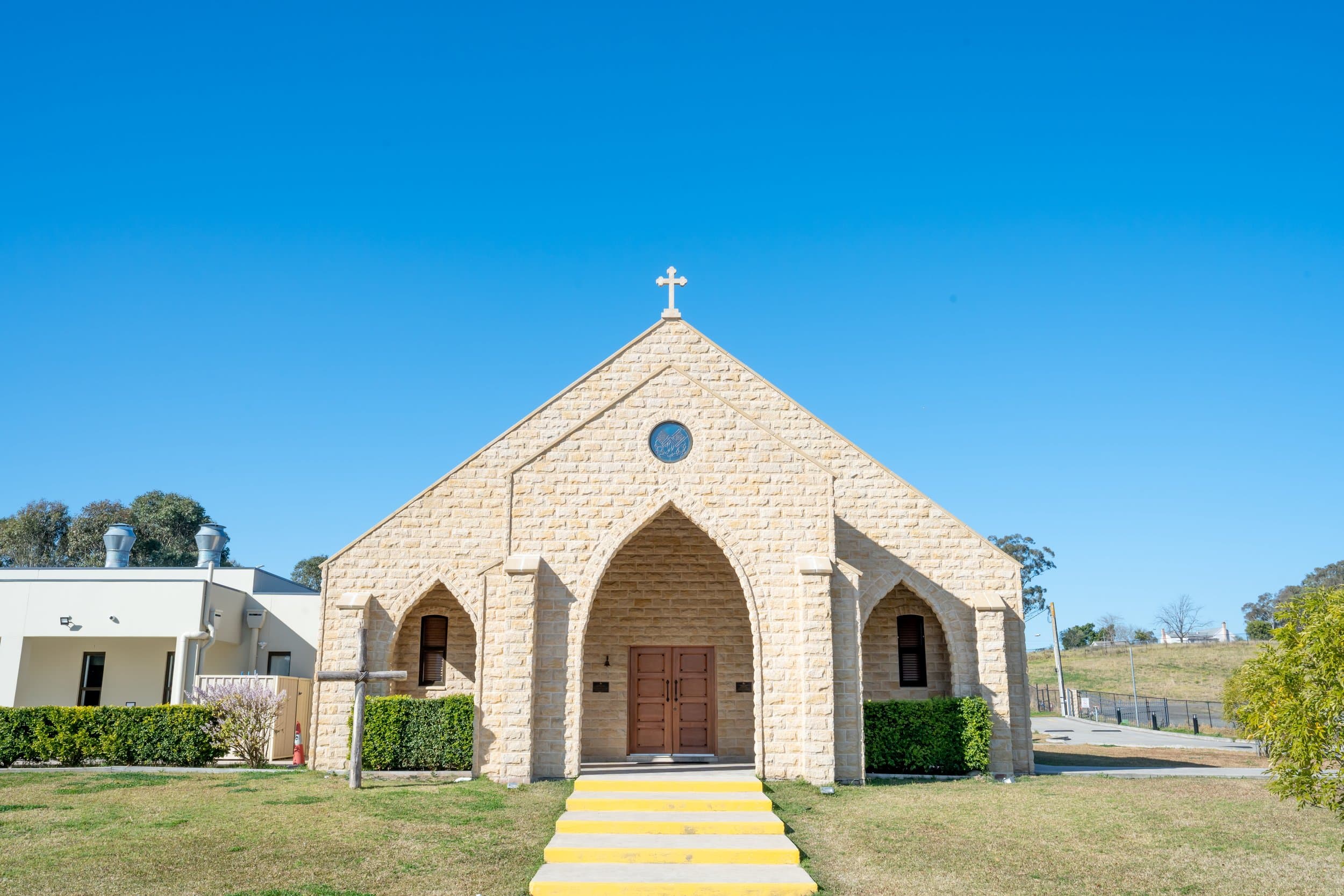 The stone gabled facade of Saints Peter and Paul Assyrian Church of the East in Cecil Park, with three pointed arches, yellow entrance steps, and a wooden cross on the lawn